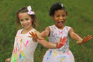 Two smiling girls covered in colorful paint hold out their messy hands together in a green grassy field.