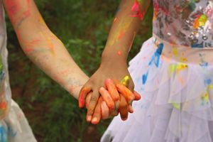Close-up of two children holding hands, their fingers and arms covered in bright orange, red and blue paint.