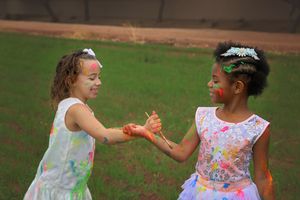 Two girls in white dresses and flower crowns hold hands and laugh while covered in colorful paint on a grassy lawn.