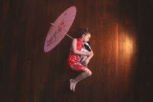 A young girl in a red qipao lies curled on a polished wood floor, holding a pink paper parasol and a stuffed toy.