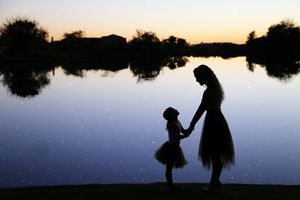 A mother and daughter in tutu skirts hold hands in silhouette beside a calm lake reflecting a colorful sunset sky.