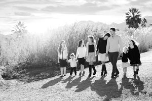 A family of eight walks hand in hand along a dirt path in black and white with palm trees and mountains behind them.