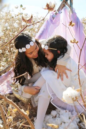 Two smiling girls in white flower crowns hug inside a pink tent surrounded by blooming cotton plants.