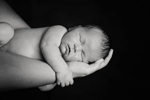 A black and white newborn portrait of a sleeping baby cradled in a parent's arms against a dark background.