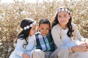 Three smiling children wearing white floral headbands sit close together in a sunny cotton field.