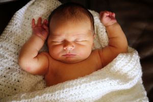 A sleeping newborn baby with raised arms rests peacefully wrapped in a soft white knit blanket.
