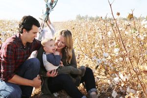 A smiling mother and father hold their baby boy while sitting among cotton plants in a golden field.