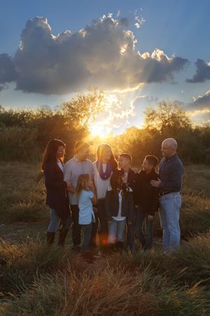 A large family gathers together in a grassy field as the sun bursts through dramatic clouds at sunset.