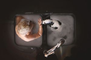 Overhead view of a person bathing a small black and white dog in a soapy double kitchen sink.