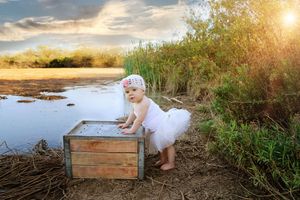 Baby girl in a white tutu and headband stands beside a wooden crate near a calm pond at golden sunset.