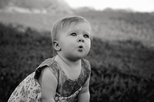 Black and white portrait of a baby girl in a floral dress puffing her lips while gazing upward outdoors.