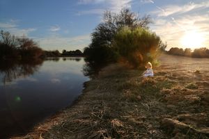 Small child in a white dress sits on a grassy pond bank as the sun sets behind trees and glowing clouds.