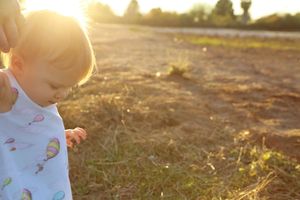 Sunlit close-up of a toddler in a balloon-print shirt walking on dry grass with a parent's hand nearby.