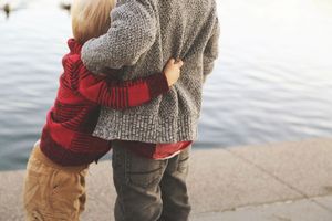 Two children in sweaters hug each other beside the water, seen from behind, in a tender sibling moment at the lakeside.