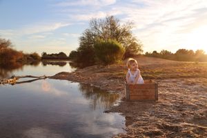Toddler girl sits on a wooden crate at the edge of a reflective pond with a warm sunset sky behind her.