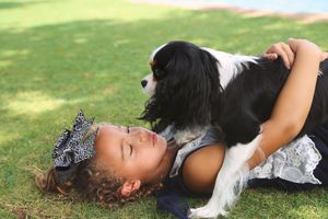 A young girl with a decorative hair bow lies on the grass with her eyes closed, hugging a black and white Cavalier King Charles Spaniel.