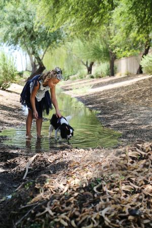 A little girl in a ruffled dress stands barefoot in a shallow stream and pets her Cavalier King Charles Spaniel among shady trees.