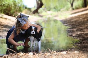 A girl in a polka dot hair bow crouches beside a shallow stream, gently hugging her black and white spaniel among the trees.