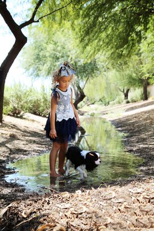 A smiling girl with a bow in her hair stands barefoot in a shallow stream next to her black and white spaniel on a sunny day.