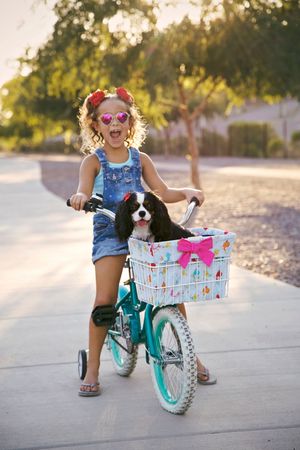 A laughing girl in heart-shaped sunglasses and a denim romper stands with her teal bike as a spaniel sits in the pink-bowed basket.