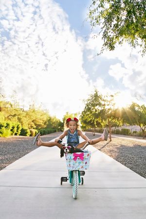A joyful girl rides a bike with her legs stretched wide, her spaniel in the pink-bowed basket, on a sunny path under a cloudy sky.