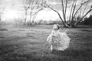 Black and white image of a little girl in a ruffled dress holding a lace parasol in a grassy field with bare trees.