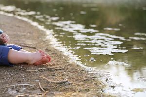 Close-up of a child's bare feet resting on a muddy shoreline beside rippling pond water.