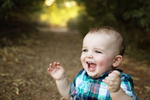 Happy baby boy in a plaid shirt laughing with hands raised on a sunlit wooded path.