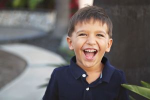 Smiling young boy in a navy polo shirt laughing joyfully outdoors beside a green plant.