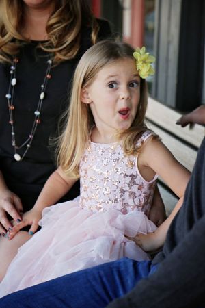 A little girl in a sparkly pink tutu dress with a yellow flower in her hair makes a surprised face while sitting with family.
