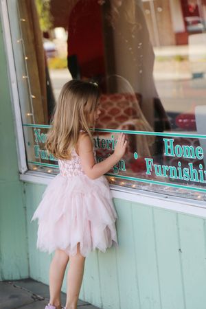 A little girl in a pink tulle dress presses her hand against a shop window with twinkle lights and home furnishings lettering.