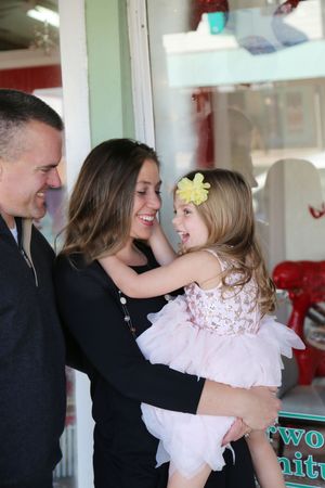 A smiling mother holds her laughing young daughter in a pink tutu dress with a yellow flower clip beside a smiling father.