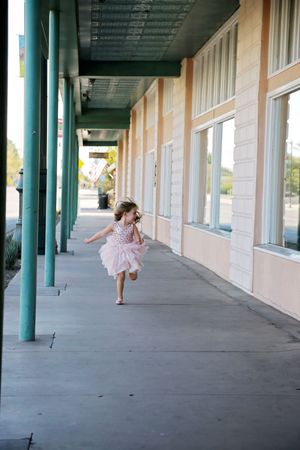 A little girl in a pink tutu dress runs joyfully along a sunlit covered sidewalk beside storefronts with teal columns.