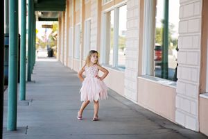 A smiling girl in a pink tulle dress stands on a covered sidewalk lined with teal pillars and storefront windows.