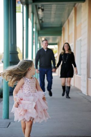 A little girl in a pink tutu dress runs down a covered sidewalk as her parents walk hand in hand behind her.