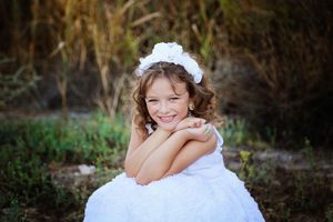 Smiling girl in a white dress and flower headband sitting with crossed arms in a grassy outdoor setting.