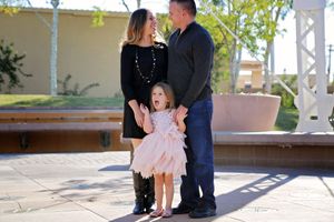 A smiling mother and father stand together outdoors on a sunny patio with their young daughter in a pink dress in front of them.