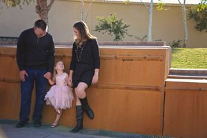 Family of three standing by a wooden wall, with a little girl in a pink tutu dress between her parents.