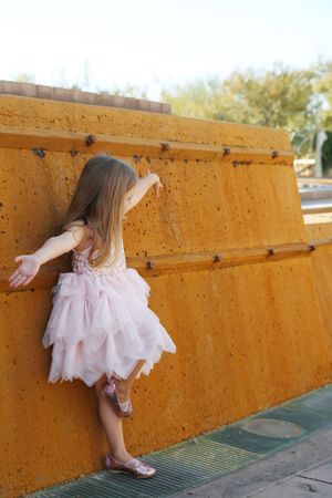 A young girl in a pink tulle dress leans against a tall orange wall with her arms outstretched and one foot raised.