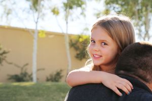 A smiling young girl with long hair rests her arms on her father's shoulder in a sunny garden with birch trees.