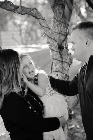 A black and white family portrait of a laughing girl in a lace dress held by her mother as her father stands beside a tree.