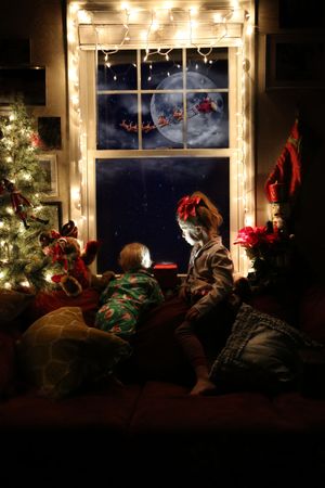 Two children in pajamas watch Santa's sleigh and reindeer cross the moon from a Christmas-lit window.