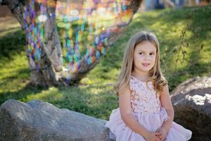 Young girl in a pink lace dress sitting on a boulder in a park with colorful sparkling bokeh behind her.