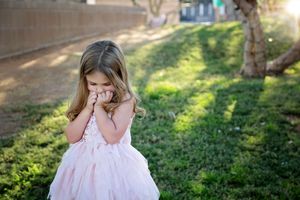 A little girl in a pink tulle dress covers her mouth with her hands while standing on sunlit grass near a tree.