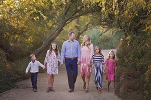 A smiling family of six walks hand in hand along a shaded dirt path beneath leafy trees during a sunny outdoor portrait session.