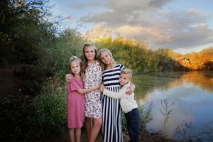 Four siblings in coordinated outfits hug and pose together beside a calm pond with golden autumn light and clouds in the background.