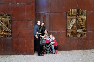 A family of five huddles together in front of a rust-colored barn wall with weathered wooden shutters in a warm outdoor portrait.