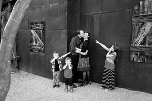 Black and white photo of parents kissing against a rustic metal wall while three young children play and point beside them.