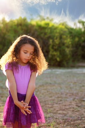 A young girl with curly hair in a purple tutu skirt stands with her hands clasped in warm golden sunlight outdoors.