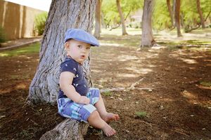 A barefoot toddler in a blue flat cap and plaid shorts sits against a tree trunk in a sunny park with dappled light.